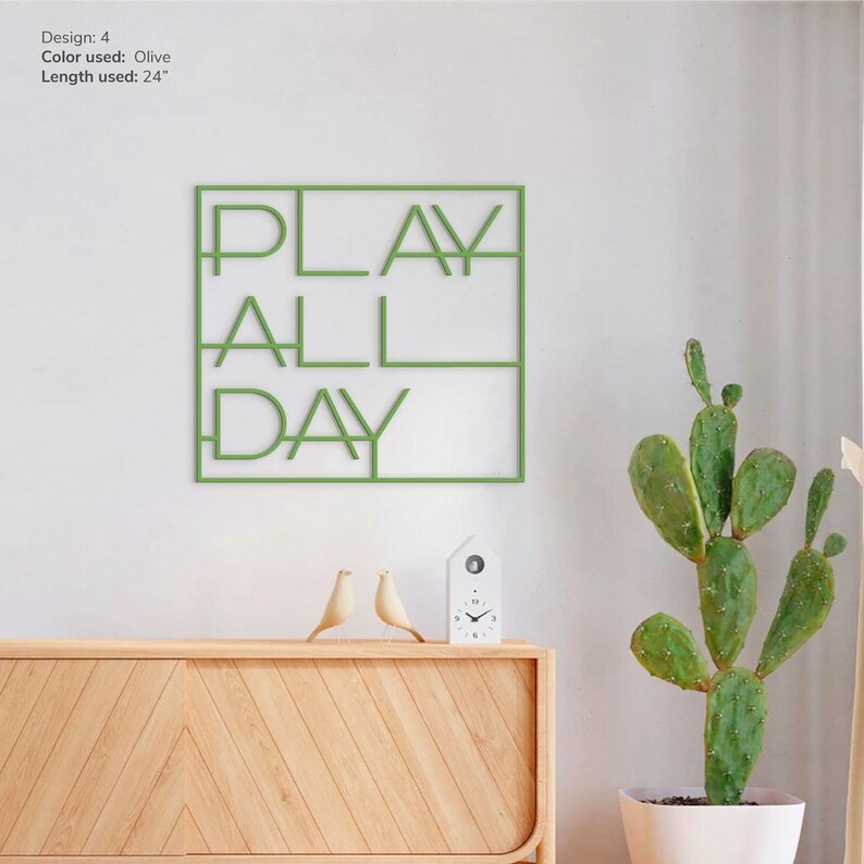 May include: Green metal wall art with the words "PLAY ALL DAY" in a modern font. The art is hanging on a white wall above a wooden cabinet.