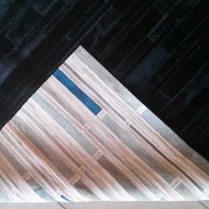 Pu&ograve; includere: Un tappeto nero con un motivo strutturato a forma di mattoni. Il tappeto &egrave; parzialmente piegato, rivelando una sezione con un design a righe nei toni del beige, grigio e blu. La trama del tappeto appare morbida.