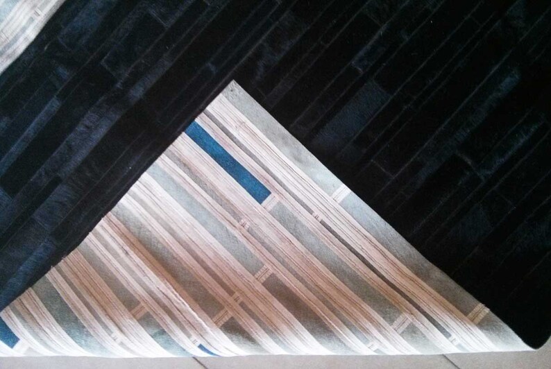 Pu&ograve; includere: Un tappeto nero con un motivo strutturato a forma di mattoni. Il tappeto &egrave; parzialmente piegato, rivelando una sezione con un design a righe nei toni del beige, grigio e blu. La trama del tappeto appare morbida.