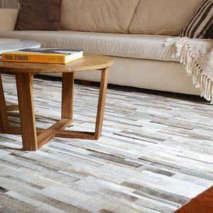 Contemporary home featuring a Taupe, Beige, Gray Striped Patchwork Cowhide Rug. Coffee tables made of wood and marble, and a soft blanket on a off-white couch.