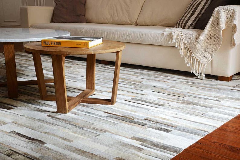 Contemporary home featuring a Taupe, Beige, Gray Striped Patchwork Cowhide Rug. Coffee tables made of wood and marble, and a soft blanket on a off-white couch.