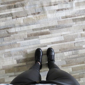 POV of a taupe striped patchwork cowhide rug. Legs in black leggings and black shoes show the scale of the stripes for this custom rug.