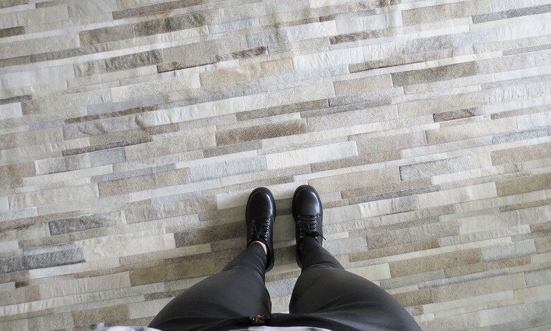 POV of a taupe striped patchwork cowhide rug. Legs in black leggings and black shoes show the scale of the stripes for this custom rug.