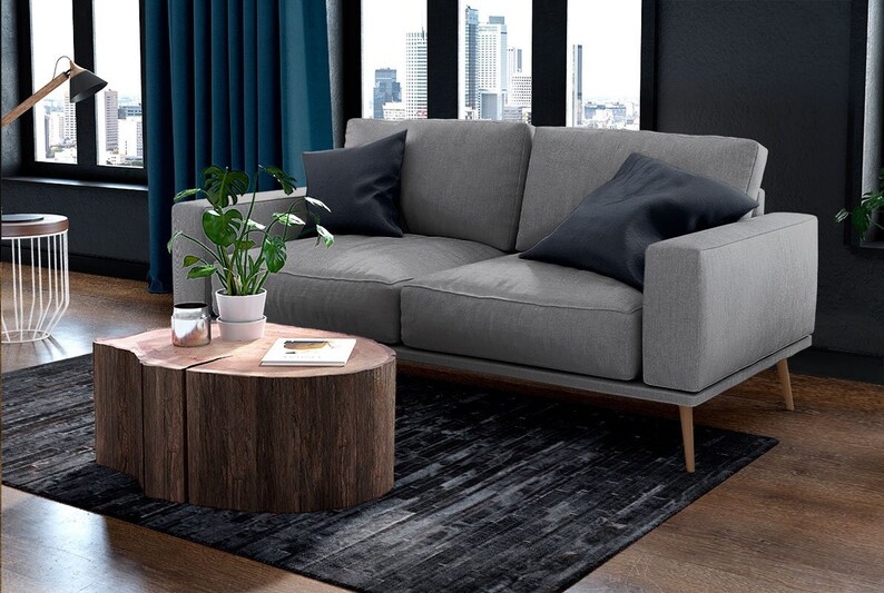 Pu&ograve; includere: Un moderno salotto con un divano grigio e due cuscini blu scuro. Un tavolino in legno, ricavato da un tronco d'albero, &egrave; posizionato di fronte al divano. Una pianta in vaso e un tappeto completano la scena.