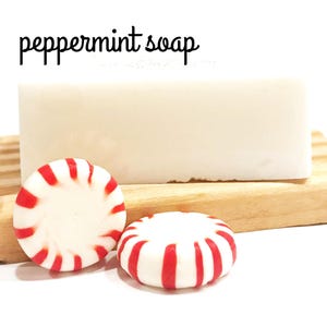 May include: A bar of white peppermint soap rests on a wooden soap dish, accompanied by two peppermint candies with red and white stripes. The text "peppermint soap" is written in black cursive above the soap.