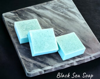 May include: Three square, light blue soaps with dark speckles are arranged on a gray, marbled tray. The soaps are labeled "Black Sea Soap" in white script.