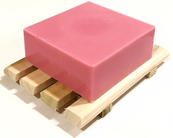 May include: A pink, rectangular bar of soap rests on a small, natural wood soap dish. The soap dish is made of several parallel wooden slats, providing drainage. The soap has a smooth surface and a solid color.