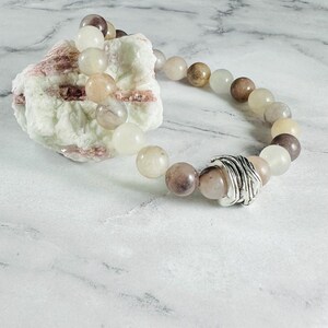 May include: A beaded bracelet featuring round, polished stones in shades of cream, beige, tan, and brown, accented by a single textured, silver-toned metal bead with a wrapped wire design, resting against a rough, pale mineral specimen.