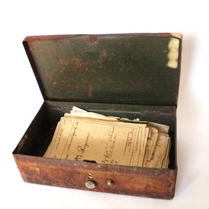May include: An open, antique metal box with a rusty brown exterior and a dark green interior. The box is filled with aged, yellowed papers. The box has a small, round latch on the front.