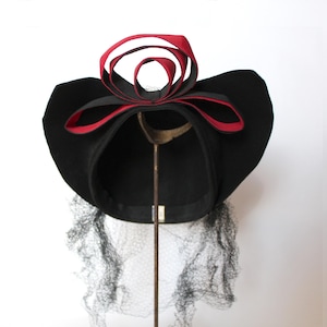 May include: A black hat with a wide brim, adorned with a red and black ribbon bow. The hat features a delicate black netting and is displayed on a metal stand. This vintage-style hat is suitable for special occasions.