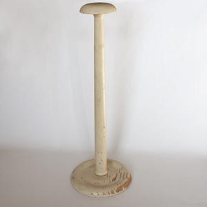 May include: A vintage off-white hat stand with a round base and a tall, slender pole. The top of the stand has a rounded shape to hold a hat. The base shows signs of wear, adding to its antique charm.
