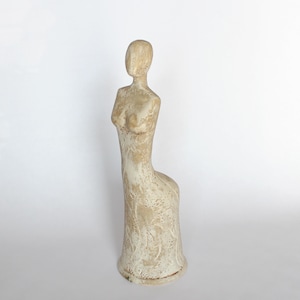 May include: A beige ceramic sculpture of a stylized female figure. The figure has a smooth, rounded head and torso, with a long, flowing dress. The sculpture has a textured surface and is standing on a round base.