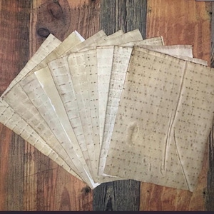 Op de afbeelding: Een verzameling rechthoekige vellen gebroken wit papier, elk voorzien van een raster van kleine, donkerbruine geperforeerde stippen. Het papier heeft een verweerd, verouderd uiterlijk met subtiele bruine vlekken en zichtbare vouwen.