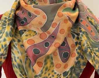 May include: A silk scarf with a vibrant abstract design. The scarf displays orange, burgundy, and grey shapes with black outlines on a tan backdrop. The edges have a yellow and green tie-dye pattern. The scarf is draped over a red top.