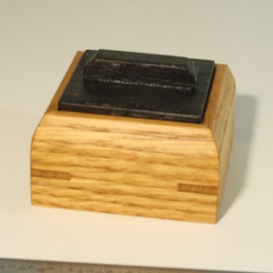 Op de afbeelding: Een kleine houten doos met een donkerhouten deksel. De doos is gemaakt van licht hout met een nerfpatroon. Het deksel heeft een verhoogde rand en een glad oppervlak.