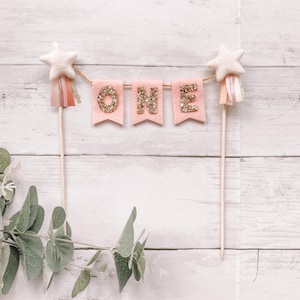 May include: Pink felt banner with gold glitter letters spelling "ONE" with white star toppers on wooden sticks.