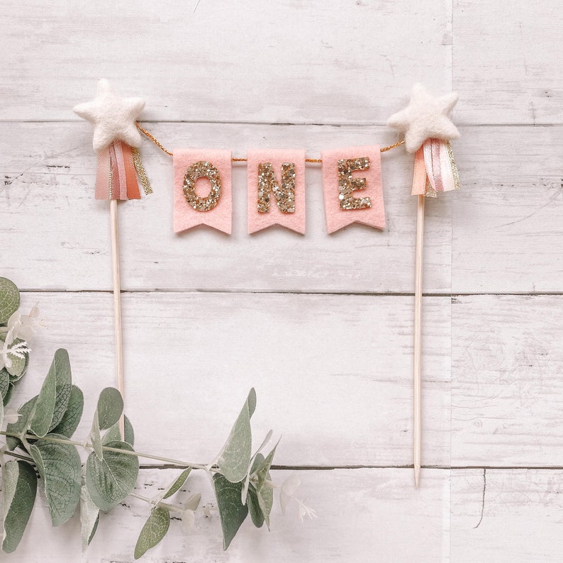 May include: Pink felt banner with gold glitter letters spelling "ONE" with white star toppers on wooden sticks.