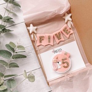 May include: Pink and gold felt cake topper with a unicorn and the word "FIVE" on a banner. The topper is in a white box with pink tissue paper.