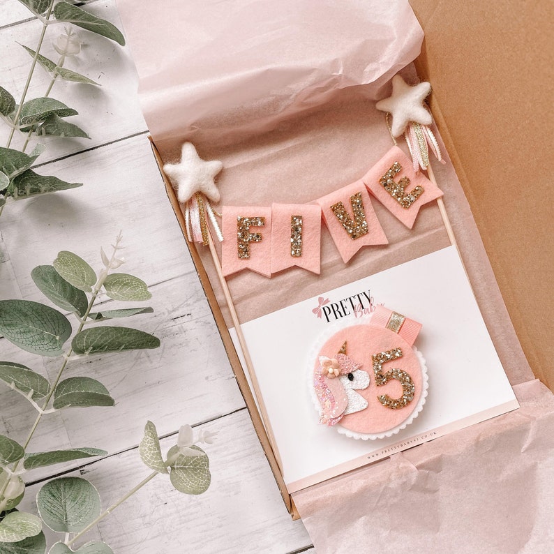 May include: Pink and gold felt cake topper with a unicorn and the word "FIVE" on a banner. The topper is in a white box with pink tissue paper.