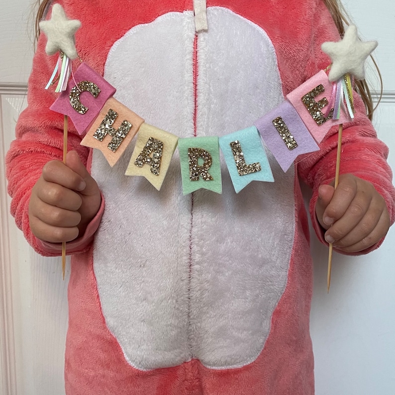 May include: A rainbow felt banner with gold glitter letters spelling "CHARLIE" with a white star and rainbow streamers on each end.