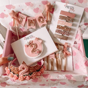 May include: A pink and gold gift box filled with a variety of items for a second birthday party. The box includes a felt banner that says "TWO", a pink and gold felt number two, a pink and gold crown, and a set of hair clips with pink and gold glitter.