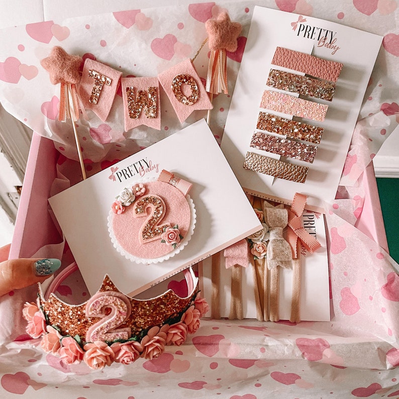 May include: A pink and gold gift box filled with a variety of items for a second birthday party. The box includes a felt banner that says "TWO", a pink and gold felt number two, a pink and gold crown, and a set of hair clips with pink and gold glitter.