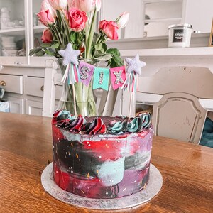 May include: A red, black, and teal birthday cake with a galaxy-like design. The cake is decorated with white sprinkles and has a topper that says "SIX" in colorful letters.