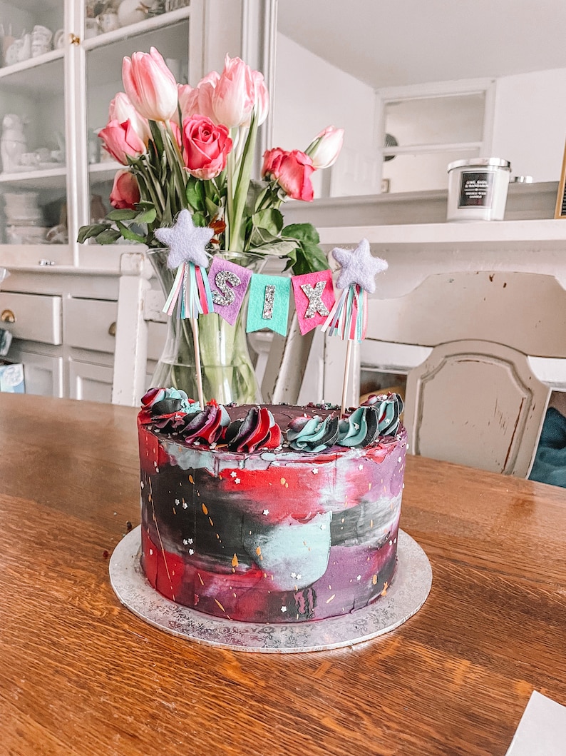 May include: A red, black, and teal birthday cake with a galaxy-like design. The cake is decorated with white sprinkles and has a topper that says "SIX" in colorful letters.