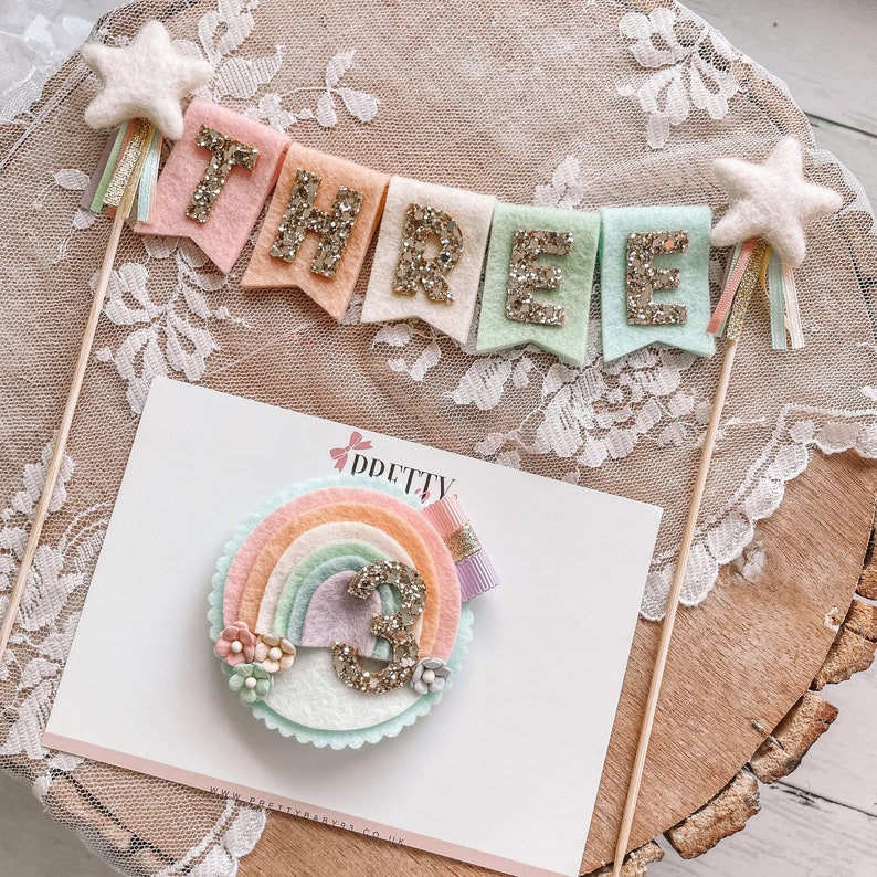 May include: A pastel rainbow cake topper with a felt number three and a banner that says "THREE" in gold glitter letters. The cake topper is perfect for a third birthday party.