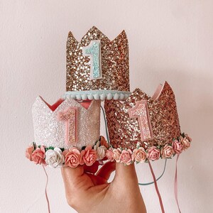 May include: Three glitter crowns with pink and white flowers and the number "1" on each. The crowns are stacked on top of each other and are held in a hand.