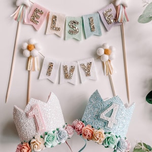 May include: Two glitter crowns with floral embellishments, one pink with the number 1 and one blue with the number 4.  Two felt banners with gold glitter letters, one says "ELSIE" and the other says "IVY".