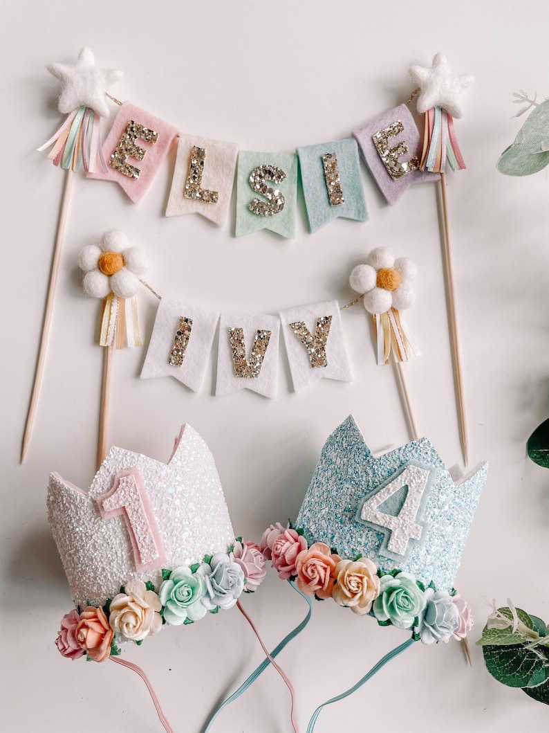 May include: Two glitter crowns with floral embellishments, one pink with the number 1 and one blue with the number 4.  Two felt banners with gold glitter letters, one says "ELSIE" and the other says "IVY".