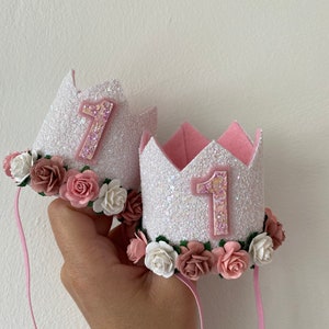 May include: Two white felt crowns with pink glitter and pink felt flowers. Each crown has a pink felt number "1" on the front. The crowns are attached to pink satin ribbons.