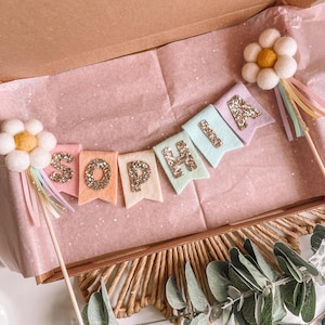 May include: A handmade banner with the name "SOPHIA" spelled out on colorful felt pennants. Each letter is embellished with gold glitter. The banner is flanked by daisy-shaped toppers with pastel ribbons, all presented in a decorative box.