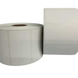 May include: Two rolls of blank white rectangular labels. One roll is partially unrolled, revealing the individual labels. The labels are on a roll with a brown cardboard core. The other roll is fully rolled.