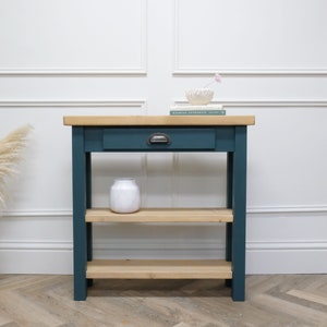 May include: A teal console table with a wooden top and two shelves. The table has a single drawer with a metal handle. A white vase and a book are on the top shelf.