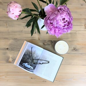 May include: A light blue vintage car on a book with a pair of glasses on top of the book. A white candle and a vase with pink peonies are on a light wood table.