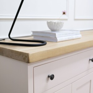 Op de afbeelding: Een lichtroze dressoir met twee lades en twee deuren. Het dressoir heeft zwarte metalen knoppen en een licht houten blad. Een zwarte lamp en een witte kom met een bloem staan op het dressoir.