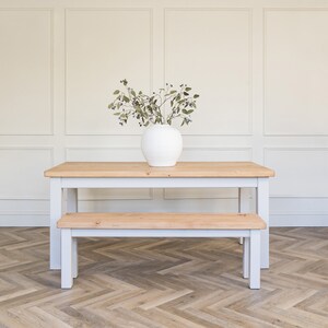 May include: A white farmhouse style table and bench set with a white vase and greenery on the table.