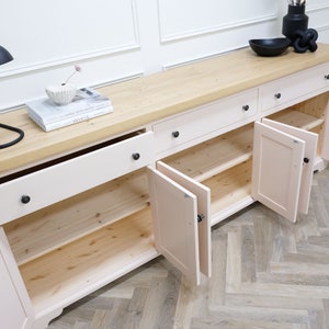 Op de afbeelding: Een lichtroze houten dressoir met vier lades en drie deuren. De deuren staan open, waardoor de binnenplanken zichtbaar zijn. De bovenkant van het dressoir is gemaakt van licht hout.