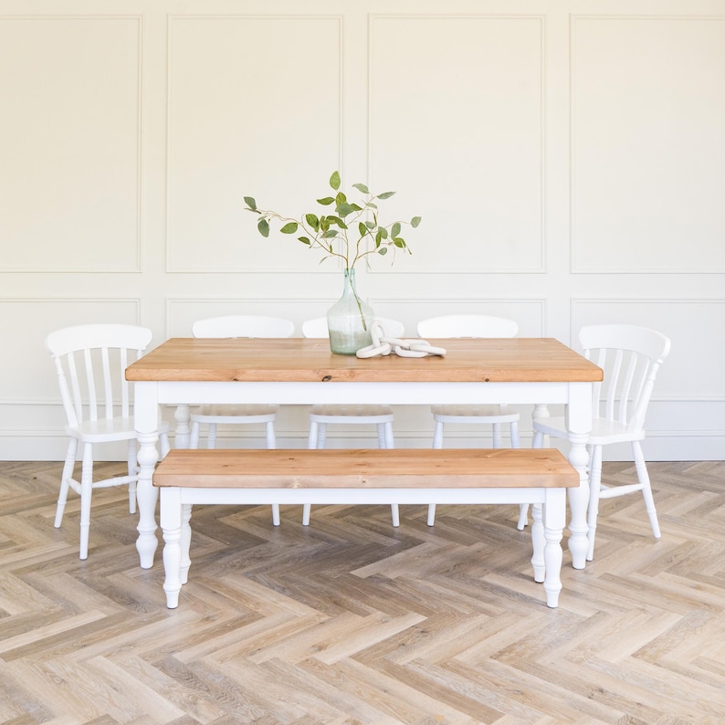 May include: A white farmhouse style dining table with a natural wood top and a matching bench. The table is set with six white chairs and a vase of greenery.