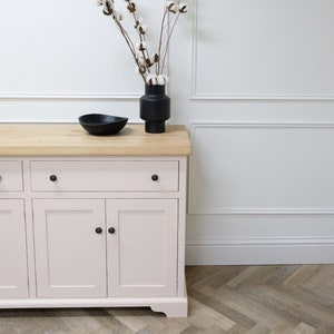 Op de afbeelding: Een lichtroze geverfde houten dressoir met drie deuren en &eacute;&eacute;n lade. Het dressoir heeft zwarte knoppen en een naturel houten blad. Een zwarte vaas met witte katoenen stelen staat op het dressoir.