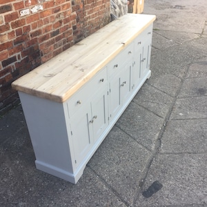 Op de afbeelding: Een grijs geverfd houten dressoir met een naturel houten blad. Het dressoir heeft zes deuren en drie laden. De deuren hebben zilveren handvatten.