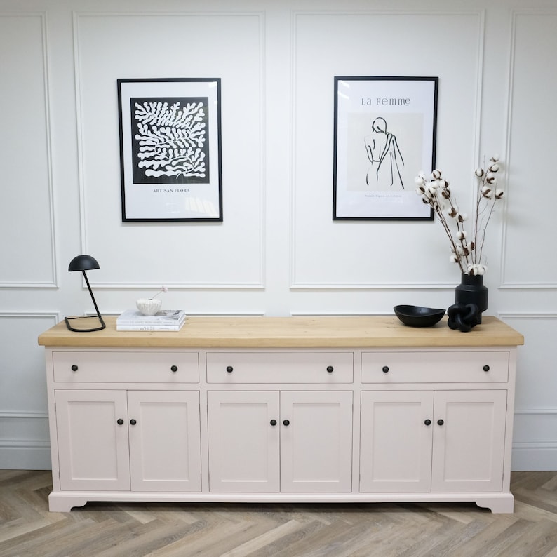 Op de afbeelding: Een lichtroze kast met zes deuren en een houten blad. De kast heeft drie lades en zes deuren met zwarte knoppen. Er hangen twee zwart-witte ingelijste prints aan de muur achter de kast.