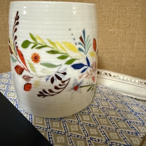 May include: A cylindrical white ceramic vase featuring a hand-painted botanical pattern of leaves and flowers in shades of green, yellow, orange, blue, and brown. The surface of the vase has subtle horizontal ridges and a smooth, glossy finish.