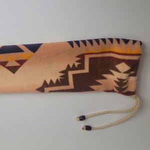 May include: A patterned fabric case in tan, brown, navy blue, and yellow, with a drawstring closure. The case has a geometric design with triangles and stepped patterns. The drawstring is cream with dark brown tips.