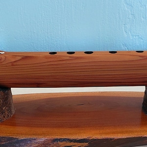 May include: A wooden flute with a carved mouthpiece and five finger holes. The flute is resting on a wooden stand.