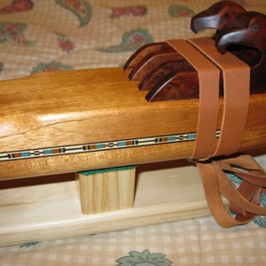 Puede incluir: Una flauta de estilo nativo americano de madera con dos cabezas de águila talladas. La flauta es de color marrón claro con una banda decorativa azul, marrón y blanca. Correas de cuero marrón aseguran las cabezas de águila. La flauta descansa sobre una base de madera.