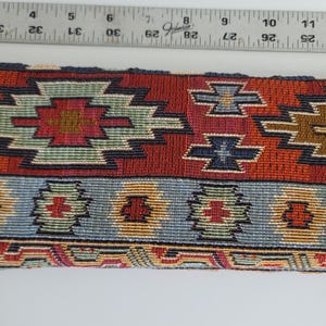 Pode incluir: Tecido tecido com um padrão geométrico em tons de vermelho, laranja, azul e castanho. O tecido tem aproximadamente 30 cm de largura, com uma régua colocada no topo para medição. O design apresenta padrões de cruz e degraus repetidos.