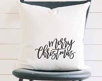 May include: A white square pillow with the words "Merry Christmas" in black script. The pillow is on a black chair against a white wooden background. The pillow is a decorative item.